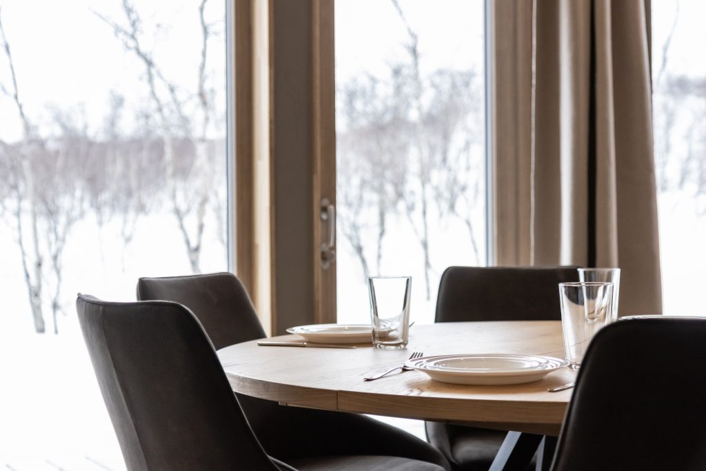 Katettu pyöreä puupöytä ja tummat tuolit suuren ikkunan ääressä, josta avautuu luminen talvimaisema